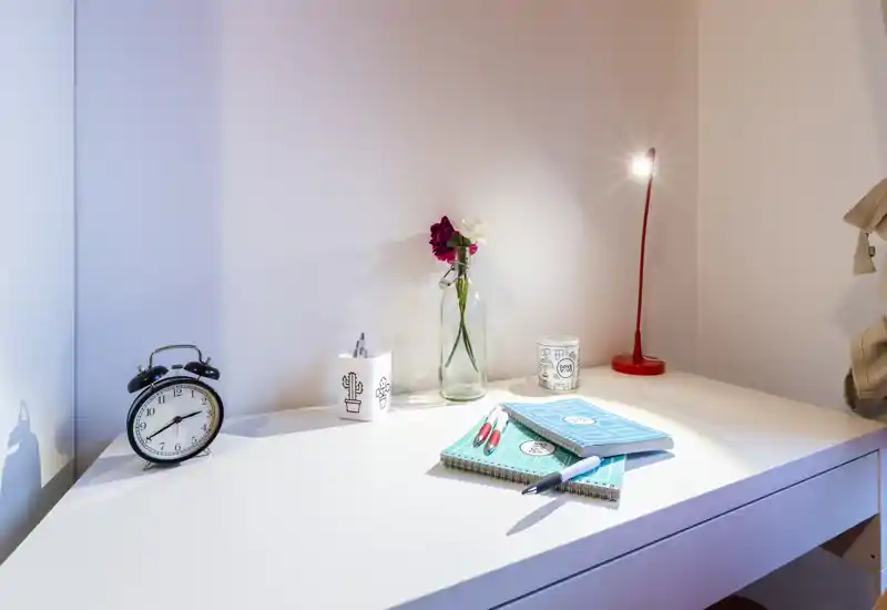Una superficie bianca ordinata da comodino o scrivania con una sveglia classica, vaso di vetro con fiori, quaderni e una sottile lampada da tavolo rossa — luminosa, minimale e dall'allestimento delicato per un angolo camera.