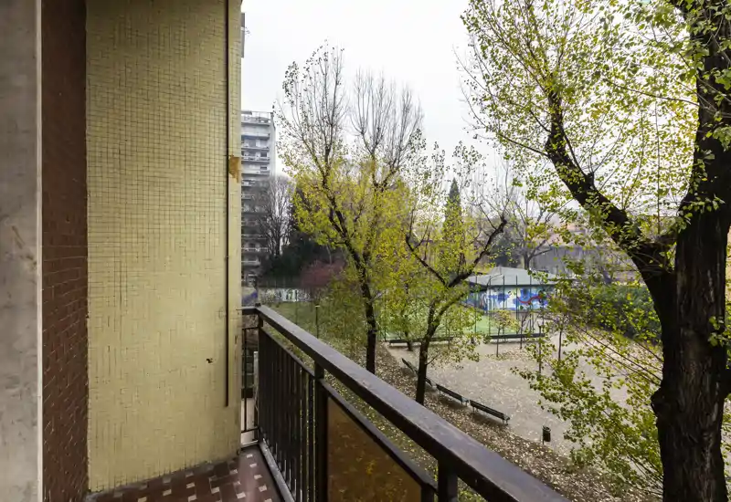 Balcone con vista su un cortile alberato e area giochi; piccolo balcone piastrellato con ringhiera in metallo e contesto urbano.