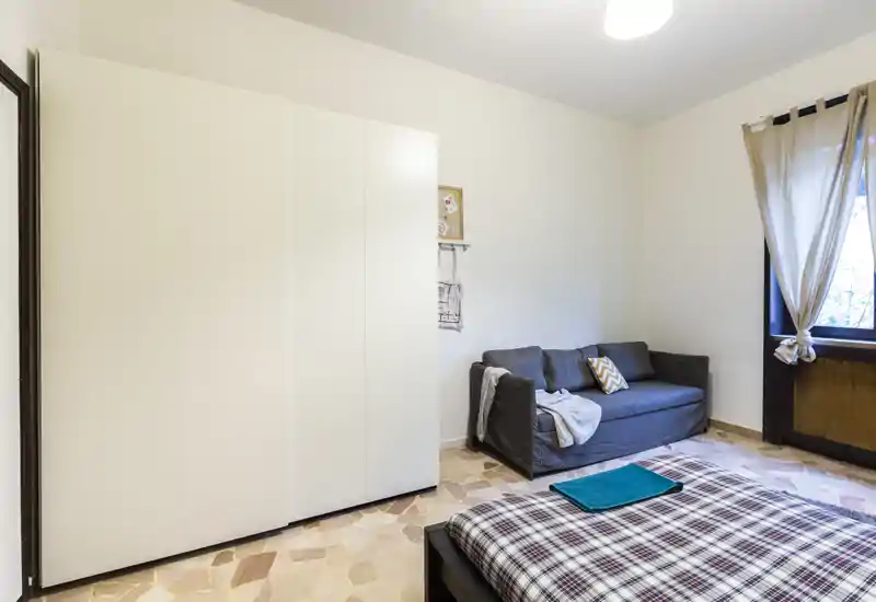 Camera da letto luminosa e semplice con letto matrimoniale in primo piano, grande armadio bianco e divano grigio vicino alla finestra. Toni neutri e luce naturale che creano un’atmosfera pulita e tranquilla, ideale per annunci.