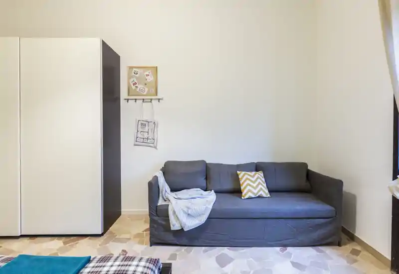 Zona giorno ben illuminata con un divano grigio compatto, cuscino decorativo e plaid, affiancata da un grande armadio — ambiente accogliente e ordinato perfetto per uno studio o un monolocale.