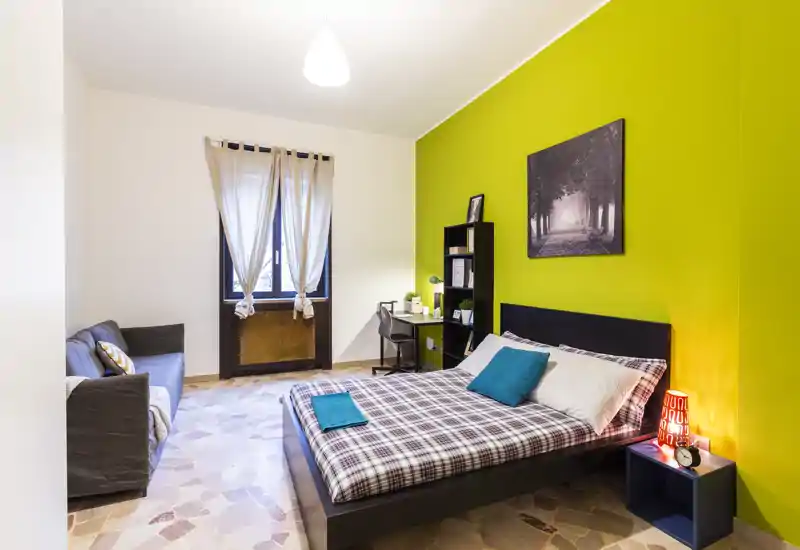 Bright, well-styled bedroom with a double bed, plaid bedding and teal accent cushions. The room features a study corner, bedside lamp and a green accent wall creating a fresh, modern atmosphere.