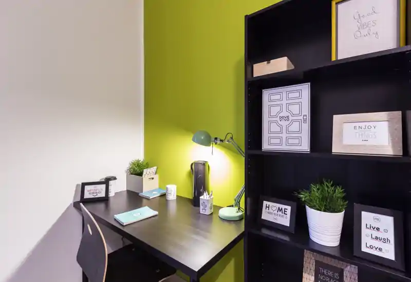 Angolo ufficio domestico luminoso e ordinato con scrivania in legno scuro, lampada verde e scaffale con cornici e piante — uno spazio di lavoro accogliente e ben organizzato.