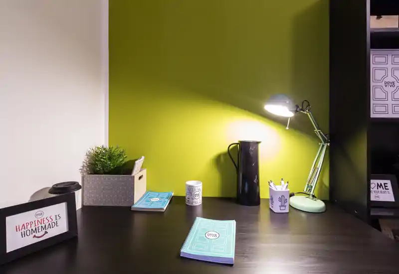 Spazio di lavoro compatto e curato con scrivania scura, parete verde e lampada da tavolo. Piccoli dettagli decorativi come pianta, quaderni e cornice lo rendono accogliente per brevi sessioni di lavoro.