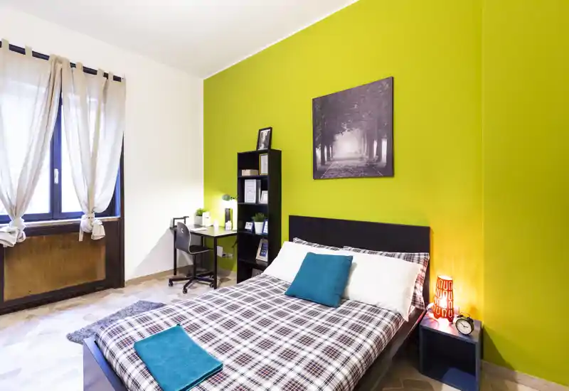 Bright, modern bedroom with a double bed dressed in plaid bedding, teal accents and a bedside lamp; a compact desk and shelving create a functional workspace next to a large window.