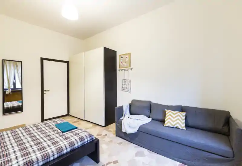 Camera da letto luminosa e semplice con letto matrimoniale, armadio e divano grigio; pavimento in piastrelle e pareti neutre creano uno spazio pulito e accogliente, ideale per brevi soggiorni.
