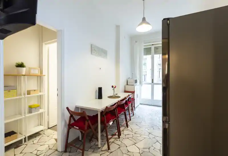 Cucina luminosa e stretta con un tavolo da pranzo e quattro sedie pieghevoli con cuscini rossi, luce naturale dalla porta del balcone e elettrodomestici moderni tra cui un frigorifero visibile.