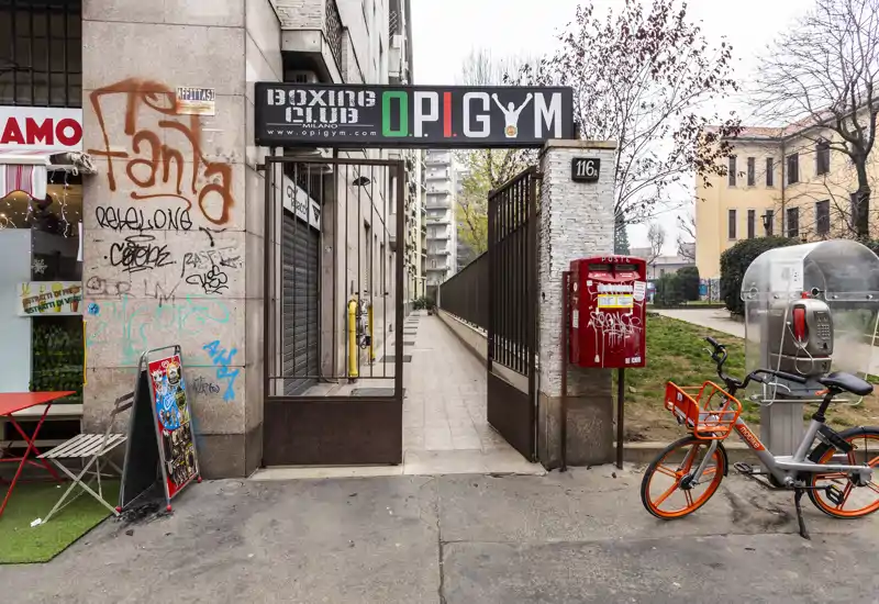 Ingresso esterno su strada con passaggio recintato verso l'edificio, dettagli urbani come una cassetta postale rossa, una bicicletta e graffiti sulla vetrina.