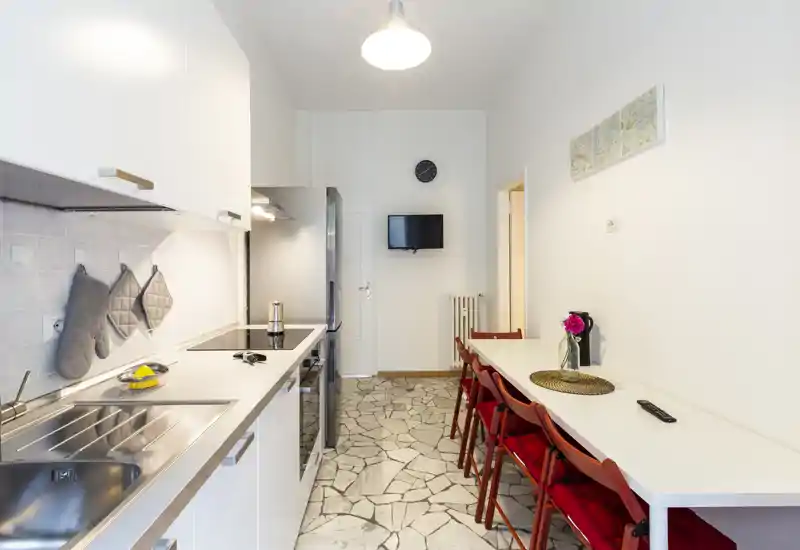 Cucina a corridoio moderna con mobili bianchi, elettrodomestici incassati e un lungo bancone pranzo con sedie rosse; luminosa, pulita e ben attrezzata per gli ospiti.