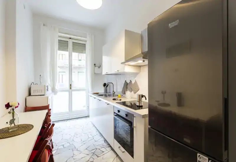Cucina moderna a corridoio, luminosa, con forno integrato, piano a induzione e frigorifero grande. La luce naturale dalla porta finestra rende l'ambiente accogliente e funzionale.