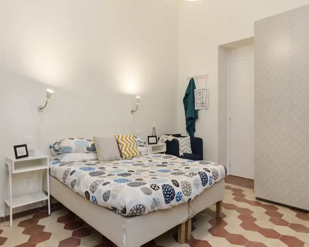 Camera da letto luminosa e accogliente con letto matrimoniale con biancheria a fantasia, comodini con applique a parete; cuscini decorativi e una poltroncina completano l'ambiente.