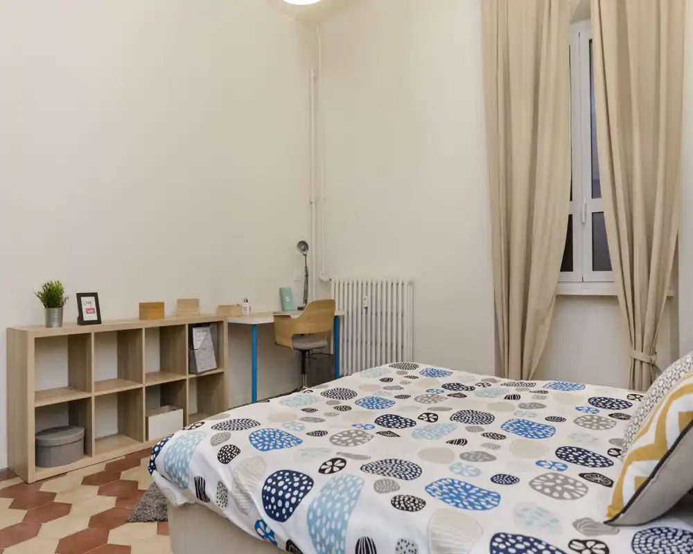 Camera da letto luminosa e ordinata con copriletto a fantasia, scrivania e scaffalatura; tende neutre e pavimento in cotto creano un’atmosfera accogliente e minimal.