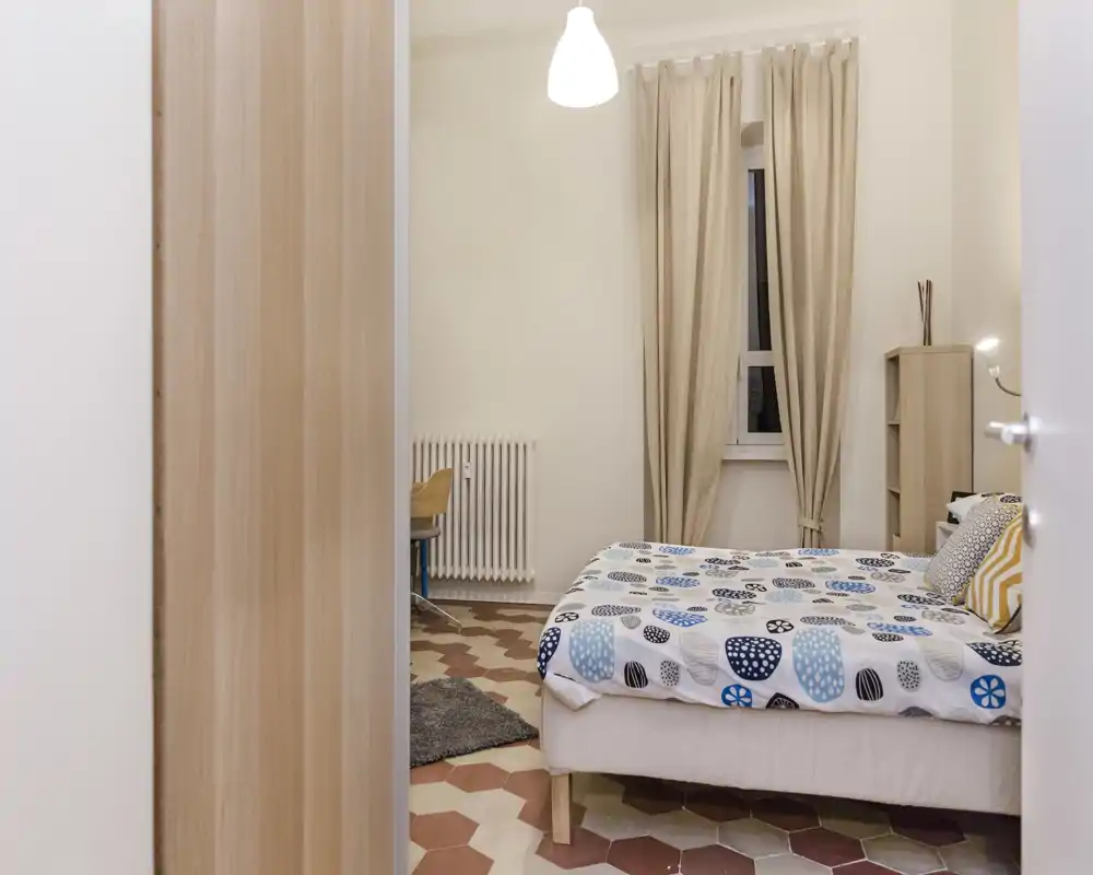 Camera da letto ben illuminata con biancheria a fantasia, tende neutre e mobili in legno semplici. Il letto è l'elemento principale e l'ambiente risulta ordinato e accogliente.
