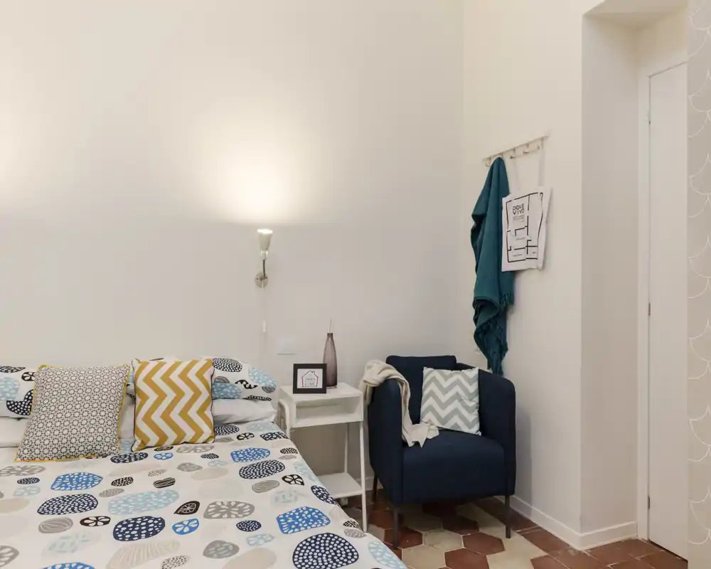 Camera da letto luminosa e ben curata con biancheria a fantasia, cuscini decorativi e una poltrona accogliente accanto al letto — perfetta per annunci di affitto che puntano su comfort e stile.