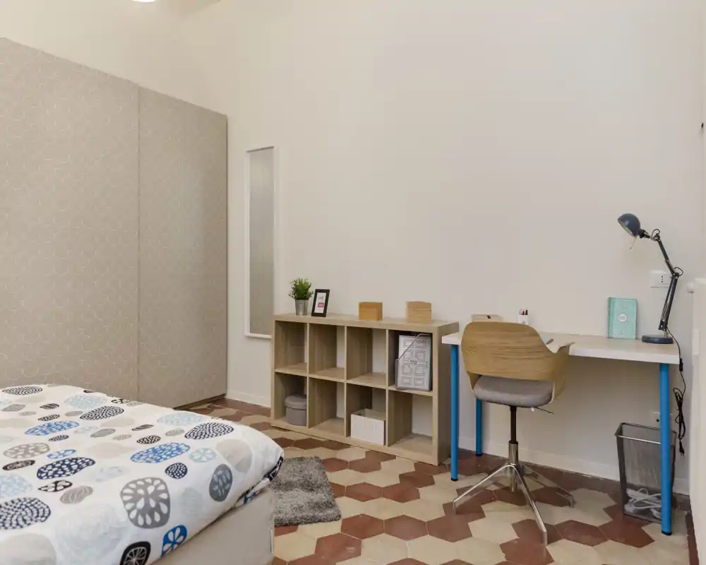 Camera da letto luminosa e ordinata con copriletto a fantasia, scaffale compatto e una scrivania semplice — ideale per una persona o uno studente.