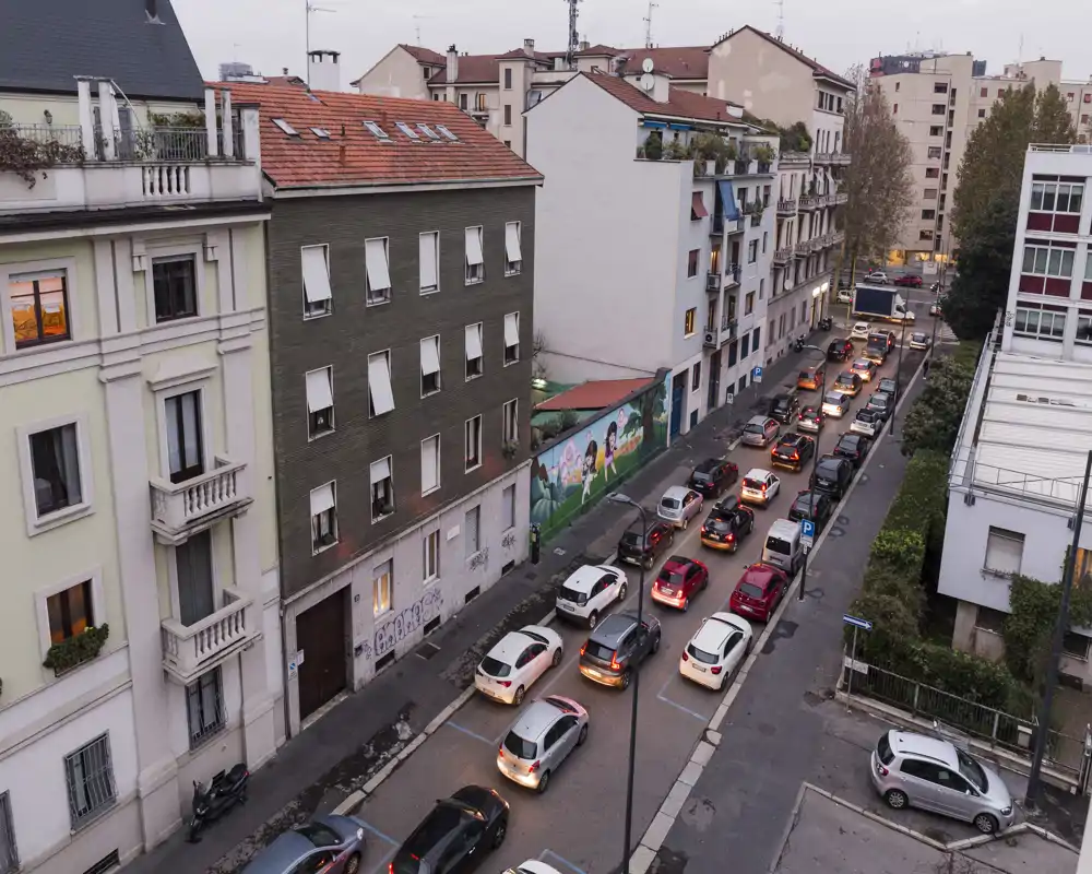 Vista urbana dall'alto con auto parcheggiate e in coda su una strada residenziale, facciate di condomini e luce serale.