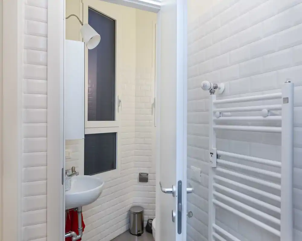 Bagno compatto e moderno con piastrelle effetto subway bianche, piccolo lavabo a muro, scaldasalviette e finestra opaca che lascia entrare luce soffusa.