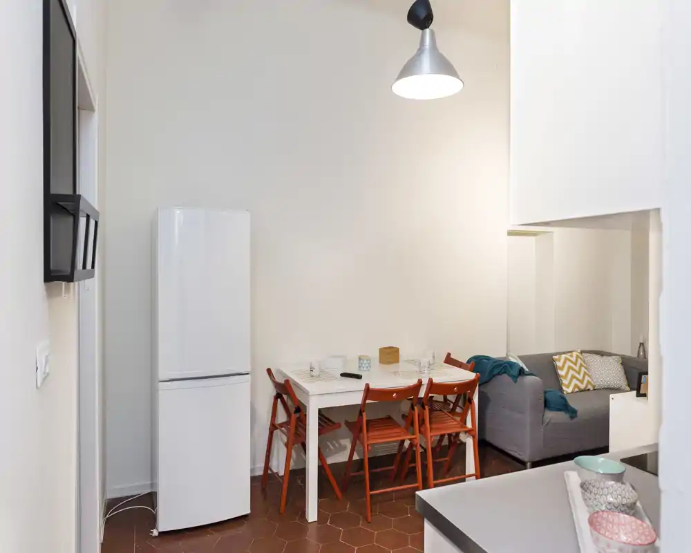 Angolo pranzo compatto con tavolo bianco e quattro sedie pieghevoli arancioni, adiacente a un piccolo divano e frigorifero — luminoso, ordinato e ideale per un appartamento in città.