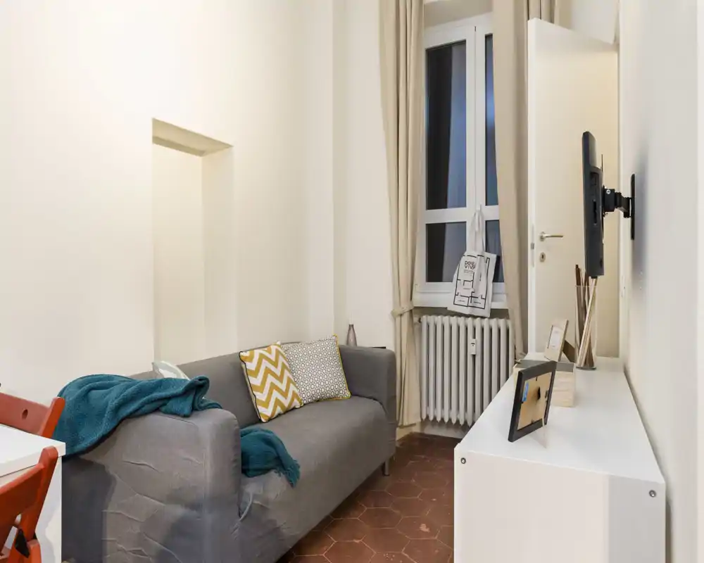 Zona living accogliente e ben illuminata con un divano grigio compatto, cuscini decorativi e TV a parete — perfetta per un soggiorno di piccole dimensioni.