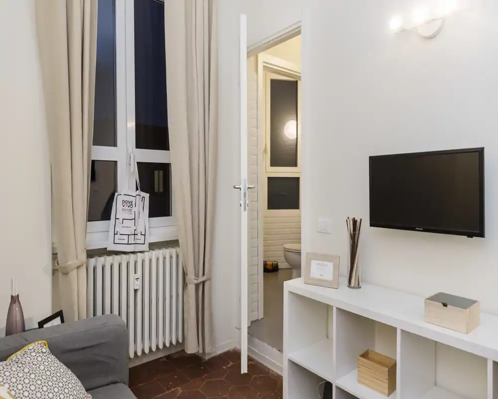 Zona giorno luminosa e ordinata con divano, TV a parete, scaffali e finestra con tende che affaccia su un piccolo bagno — angolo accogliente e moderno di un appartamento urbano.