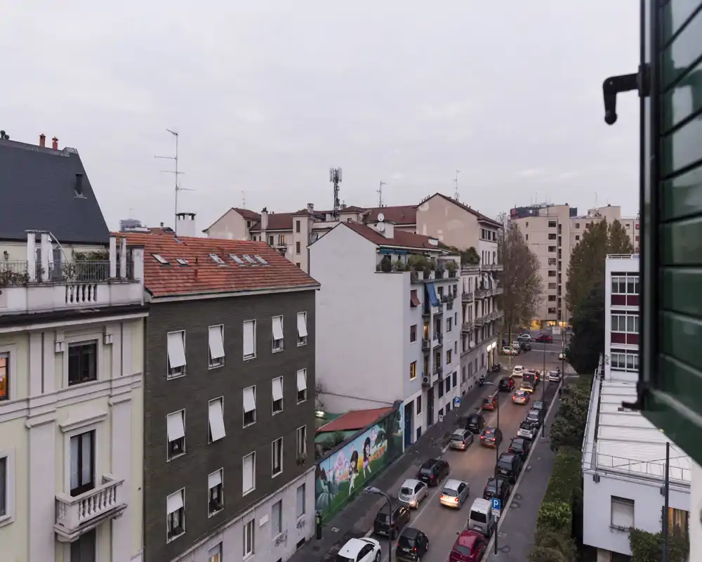 Vista sulla strada da una finestra residenziale con edifici adiacenti, auto parcheggiate e un murale urbano; luce nuvolosa neutra e dettagli architettonici nitidi.