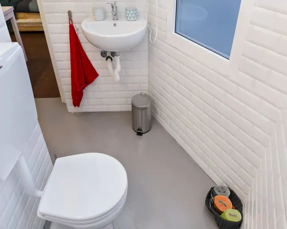 Baño compacto y limpio con lavabo de pared, inodoro y paredes alicatadas. Espacio luminoso con elementos modernos y un pequeño cubo de basura.