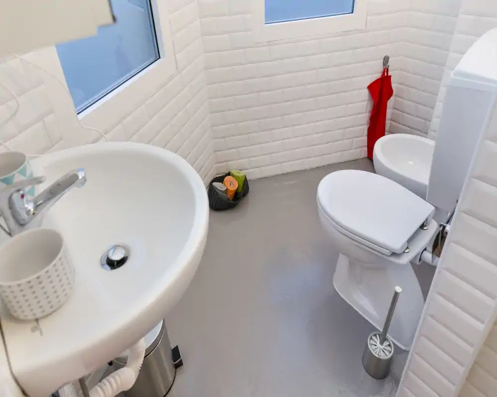 Baño compacto y limpio con paredes tipo metro blancas, lavabo, inodoro, bidé y suelo neutro—luminoso y bien cuidado.