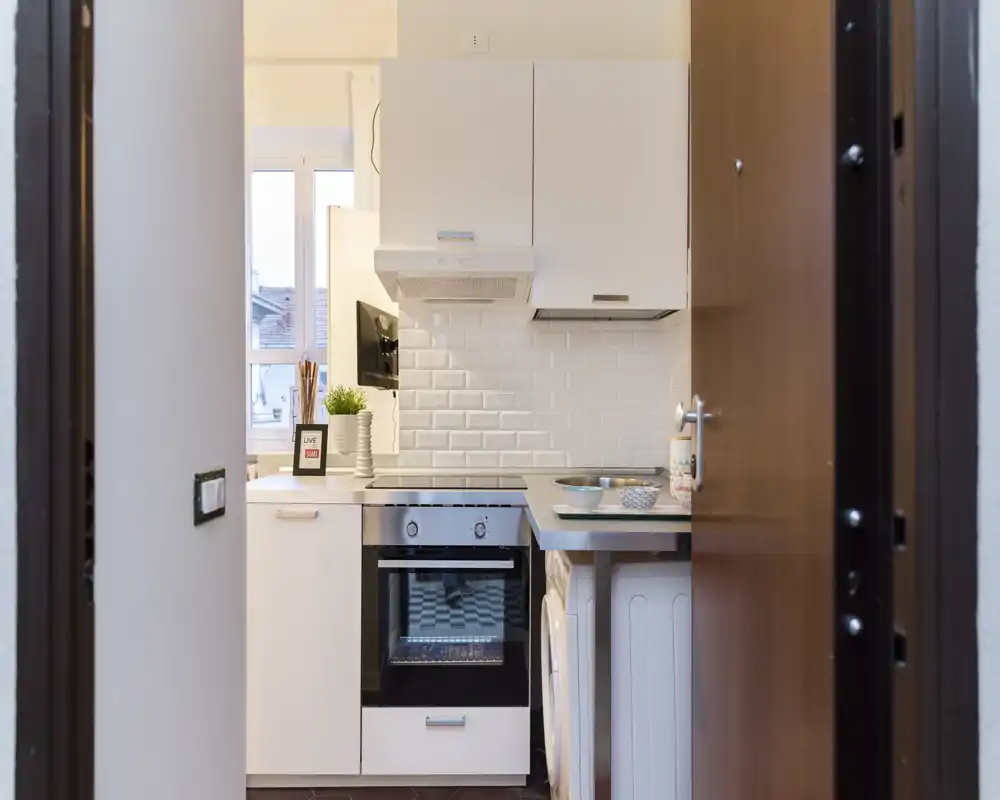 Cocina compacta y luminosa con armarios blancos, horno empotrado, salpicadero de azulejos y pequeño encimera — ideal para un apartamento urbano acogedor.