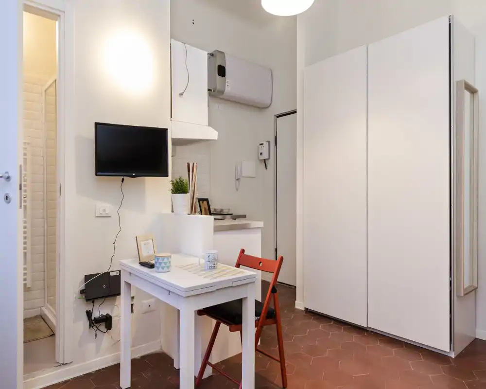 Zona de estar compacta y luminosa con mesa pequeña, silla plegable roja, televisor en la pared y gran armario blanco — distribución práctica ideal para un estudio.