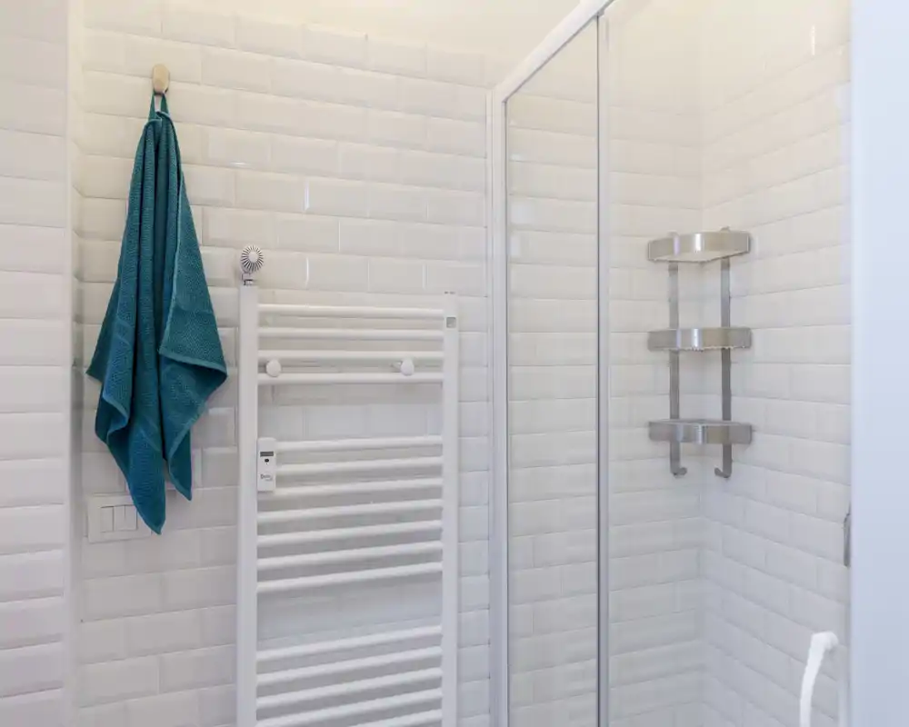 Rincón de baño moderno y luminoso con azulejos blancos tipo metro, mampara de ducha de vidrio, toallero radiador y una toalla turquesa colgada.