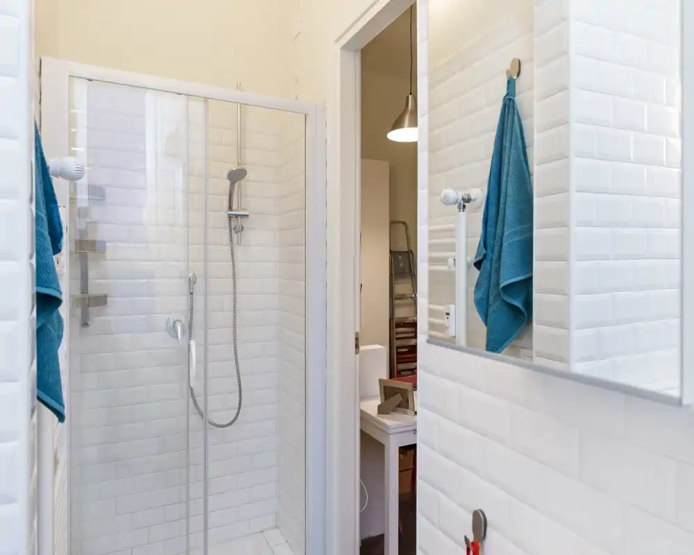 Baño moderno y luminoso con ducha acristalada, azulejos blancos tipo 'subway' y espejo en la pared; las toallas turquesa aportan color.