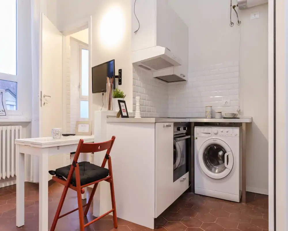 Cocina compacta y moderna con muebles blancos, horno empotrado y lavadora bajo la encimera; luminosa, ordenada y eficiente para un apartamento pequeño.