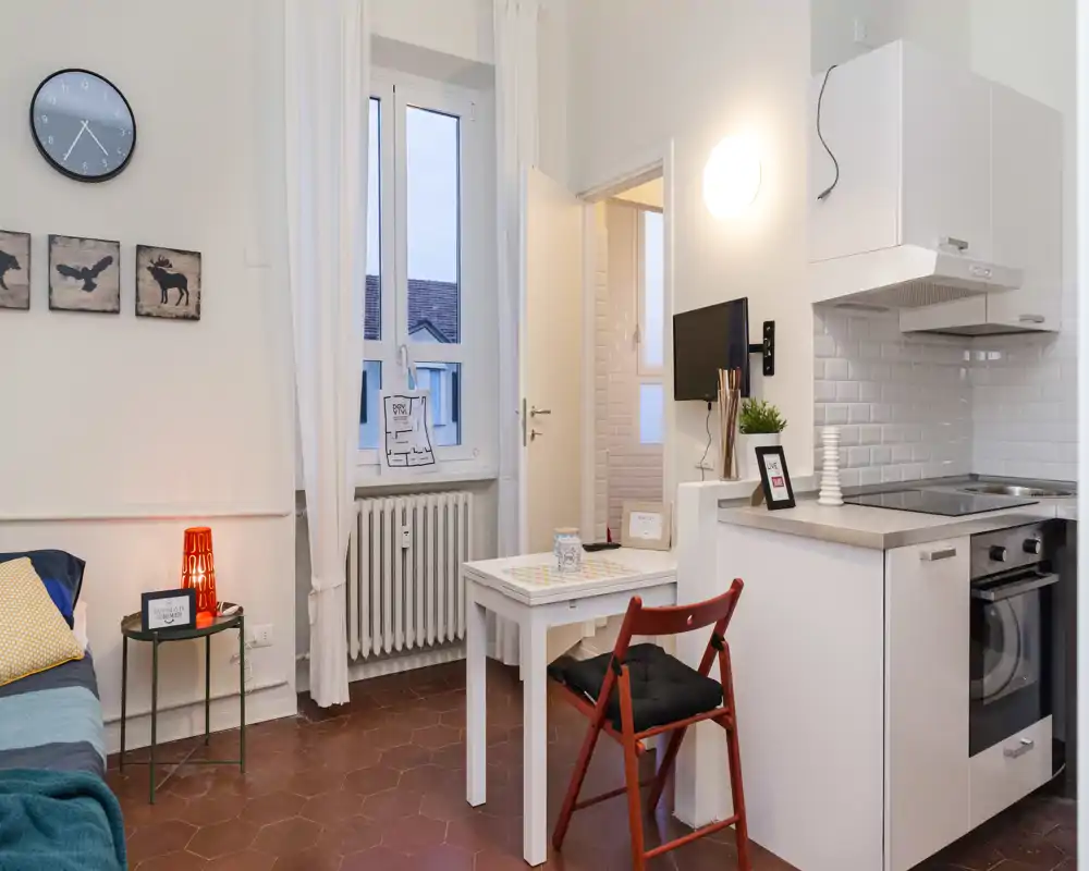 Dormitorio tipo estudio con cocina americana. Se aprecia parte de la cama a la izquierda, una mesa pequeña con silla y una cocina moderna con horno y placa — ideal para una estancia individual cómoda.