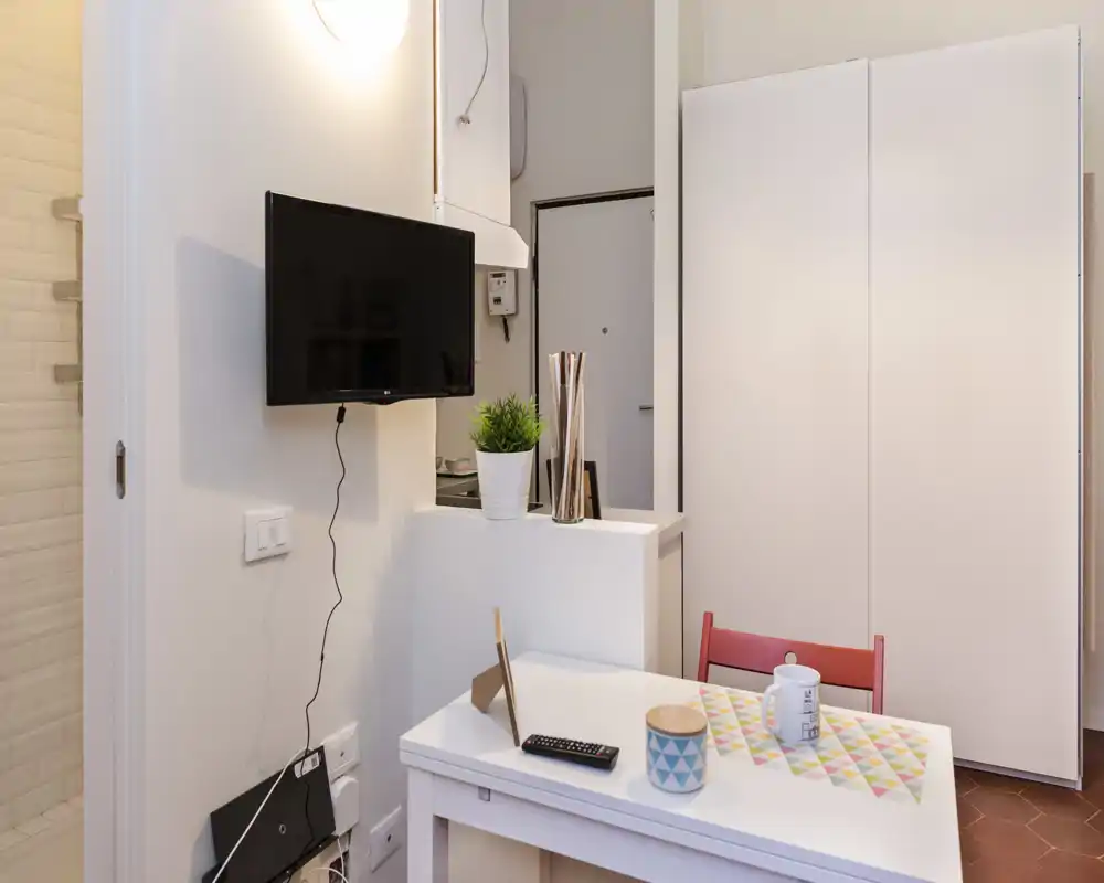 Zona de estar compacta y luminosa con mesa pequeña, televisor mural y armario; decorada con planta y menaje, ideal para un estudio.