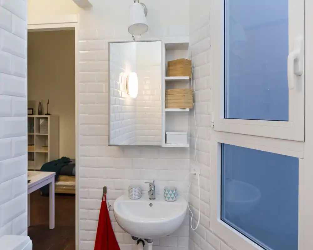 Baño compacto y moderno con azulejos blancos tipo 'subway', lavabo mural, mueble con espejo y una gran ventana translúcida que aporta luz natural.