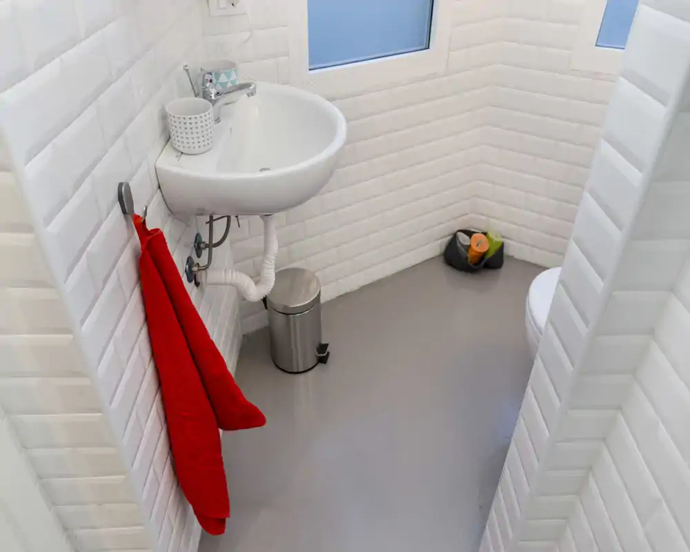 Baño compacto y moderno con lavabo mural, inodoro parcialmente visible y paredes de azulejos blancos; accesorios minimalistas y suelo brillante.