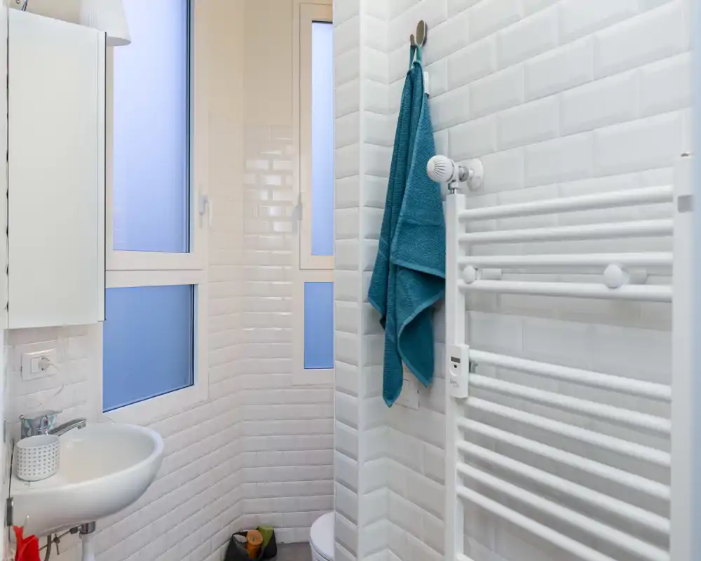 Baño compacto y moderno con azulejos tipo metro blancos, lavabo pequeño, radiador toallero y ventanas translúcidas que ofrecen privacidad y luz natural.