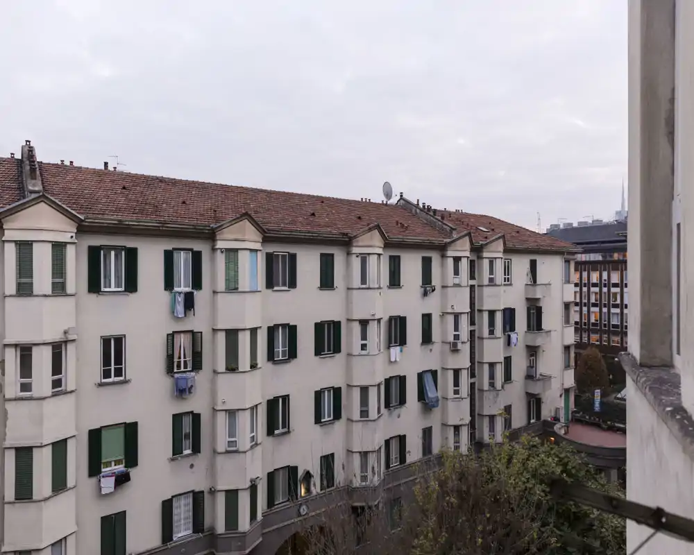 Vista de la fachada de un bloque de pisos y los tejados vecinos desde un balcón o ventana; se aprecian ventanas, contraventanas y ropa tendida con cielo nublado.