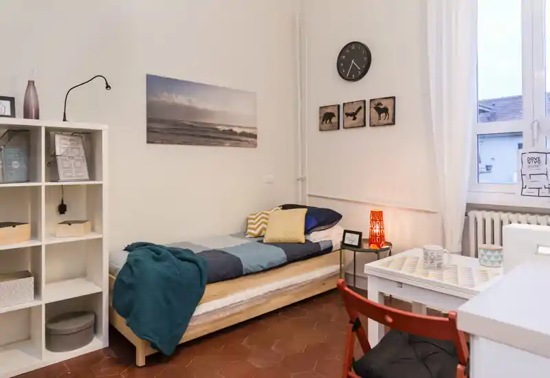 Pequeña habitación individual bien decorada con cama de madera, textiles coloridos, lámpara de noche y ventana luminosa — ideal para una estancia acogedora.