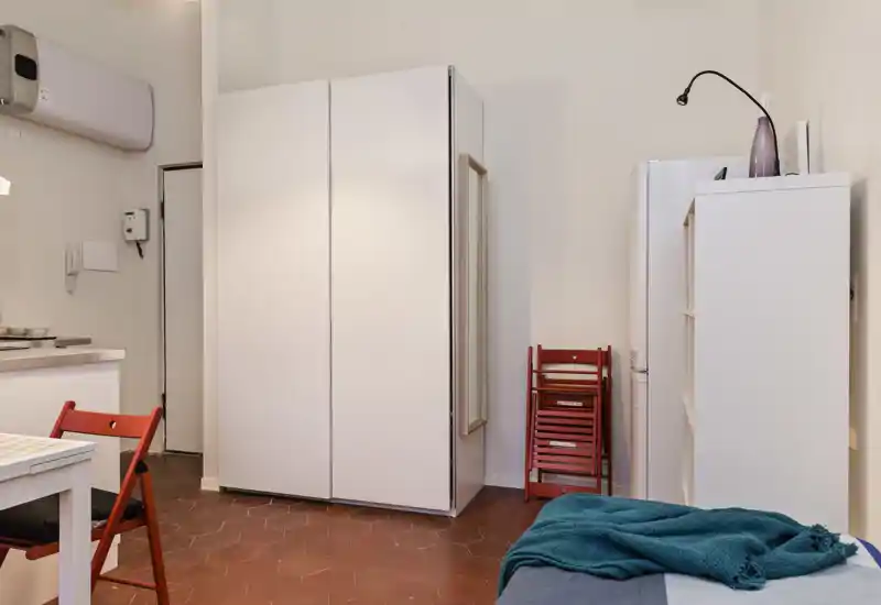 Rincón de dormitorio de estudio compacto con armario blanco, mesa pequeña y esquina de cama visible con una manta verde azulado; espacio luminoso y ordenado, perfecto para una persona.