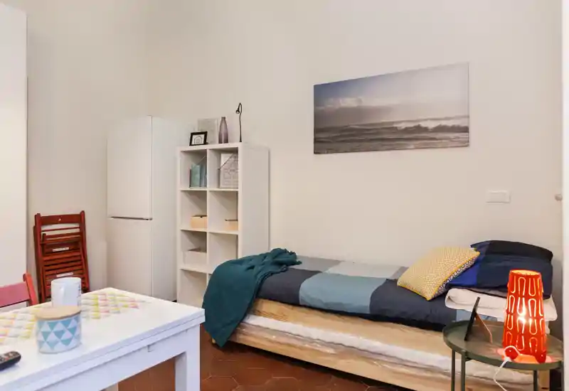 Dormitorio compacto con cama individual vestida con varias capas, manta acogedora y cojines; marco de madera clara y cuadro en la pared que crean un ambiente tranquilo.