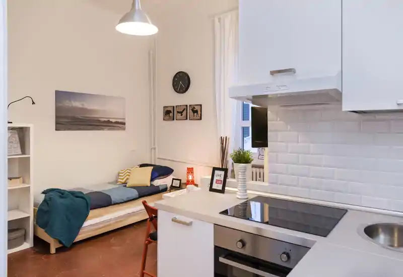 Estudio compacto con cama individual junto a una pequeña cocina abierta; tejidos cuidados, lámpara y cuadros en la pared crean un espacio acogedor y bien iluminado, ideal para estancias cortas.