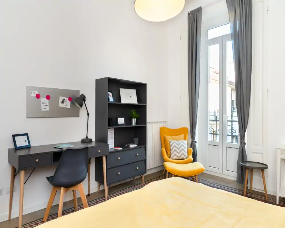 Dormitorio moderno y luminoso con la cama bien hecha en primer plano, escritorio y estantería, y un cómodo sillón amarillo junto a la puerta balcón que aporta luz natural.