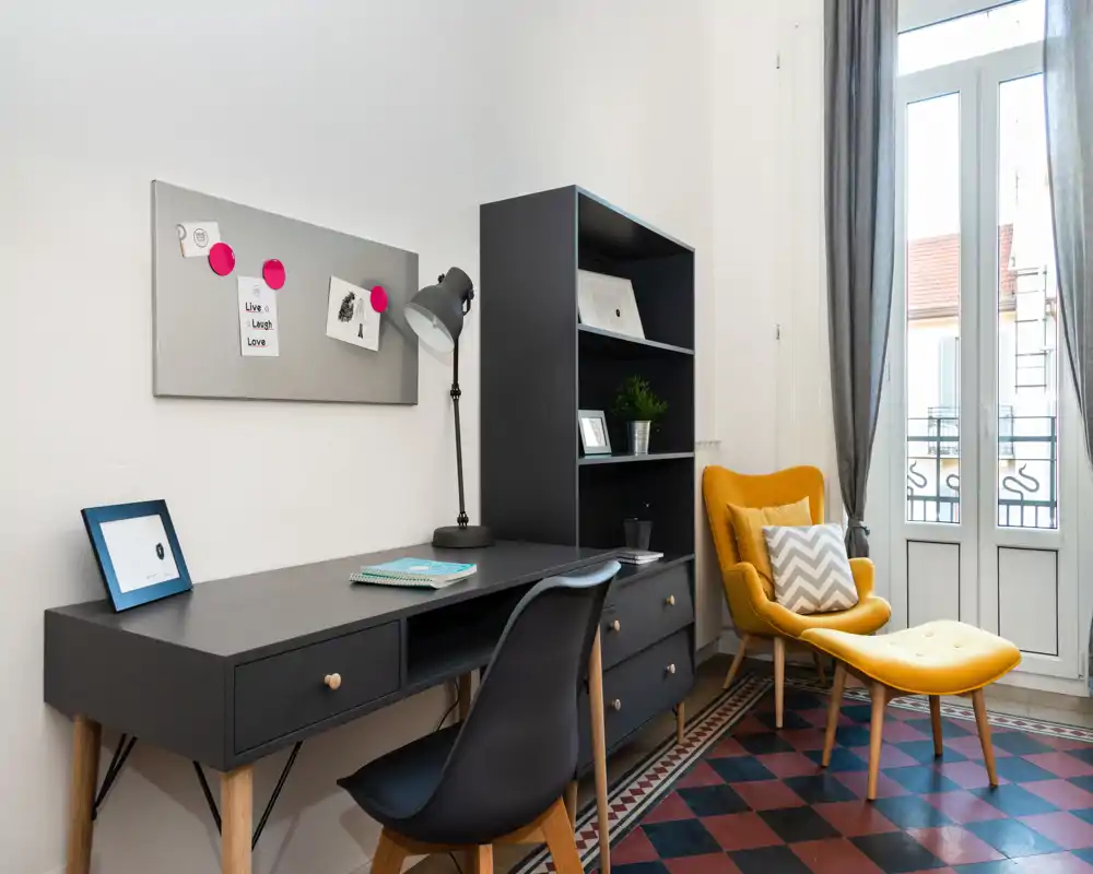 Zona de estar con estilo: escritorio y estantería oscuros, butaca amarilla con reposapiés, suelo de baldosas decorativas y ventanas altas que inundan la estancia de luz natural — perfecto como rincón de lectura o trabajo acogedor.