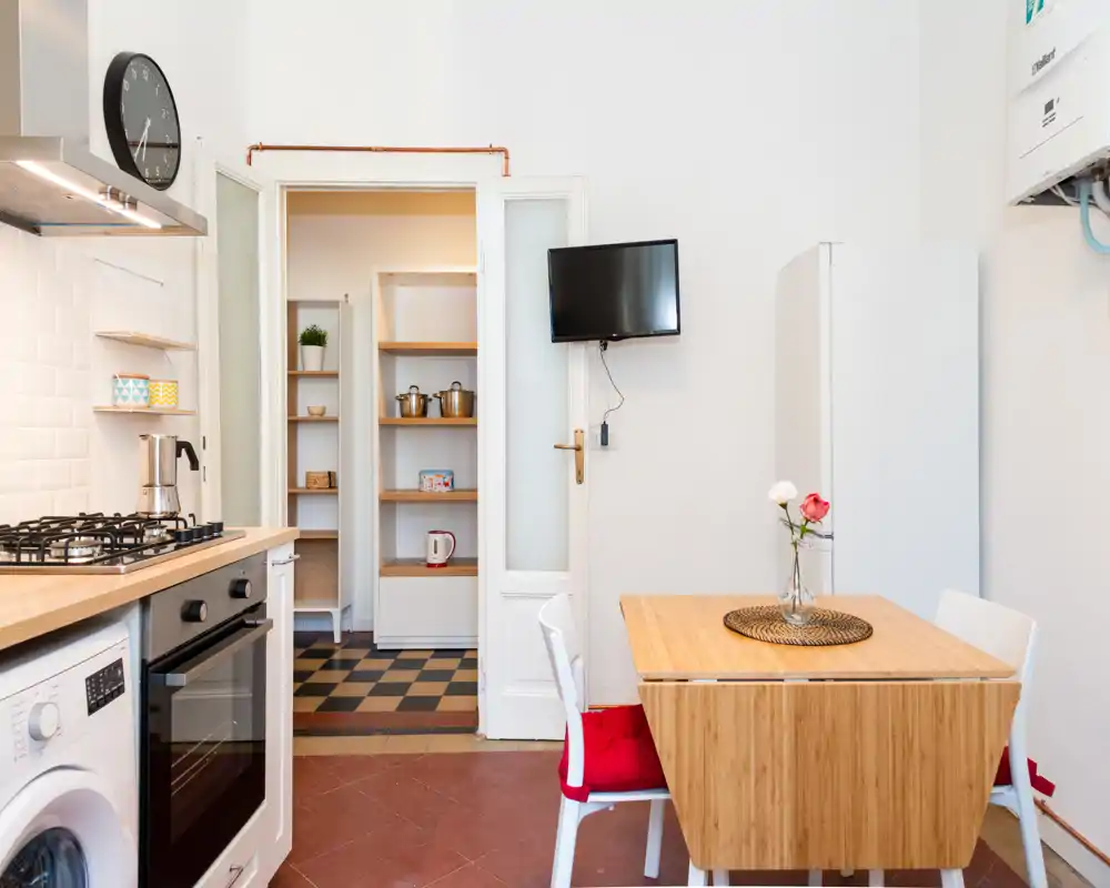 Cocina luminosa y ordenada con placa de gas, horno empotrado y mesa de comedor compacta. Las encimeras de madera y el pequeño jarrón con flores crean un espacio acogedor y funcional, ideal para estancias cortas.