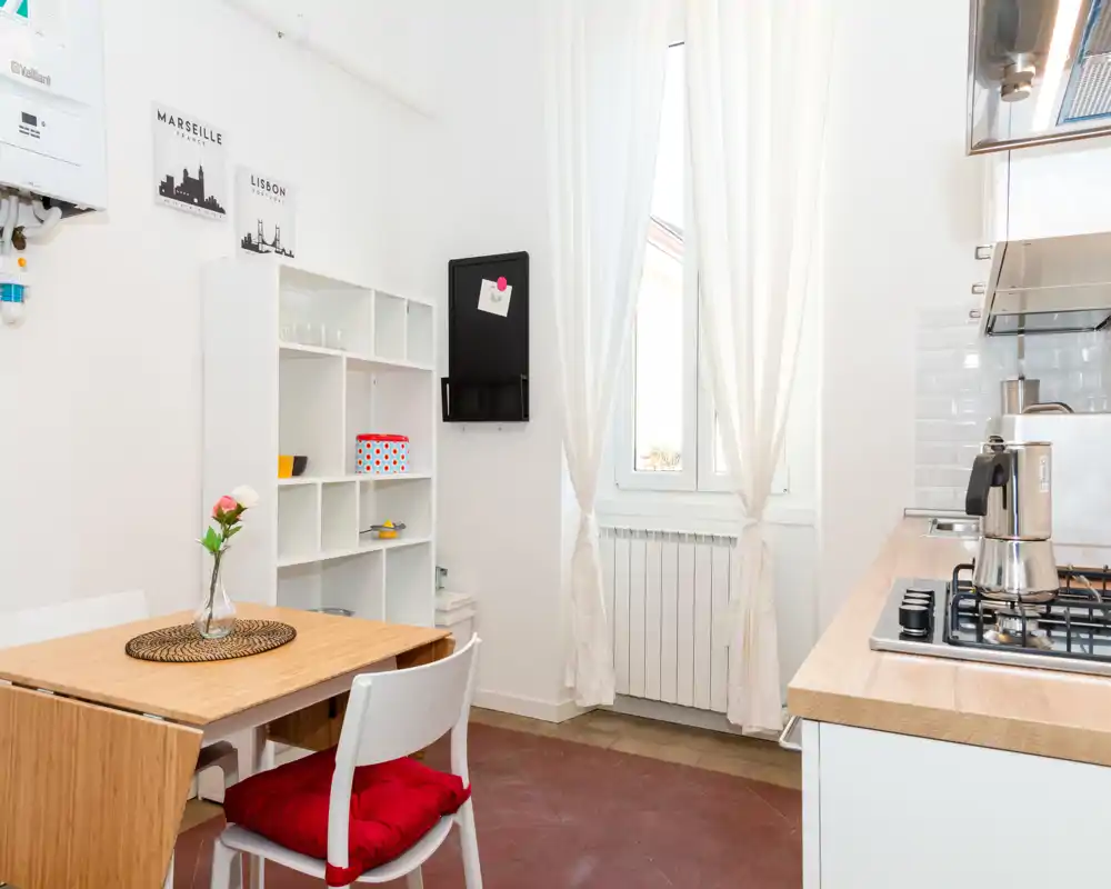 Cocina compacta y luminosa con una pequeña mesa de comedor y estanterías minimalistas. La luz natural desde la ventana realza las encimeras de madera y el rincón para desayunar.