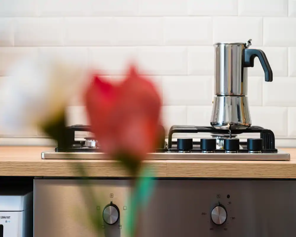 Plano cercano de una placa de cocina moderna con una cafetera moka en el fuego, encimera de madera y azulejos blancos en el fondo; una flor desenfocada en primer plano aporta un toque decorativo.