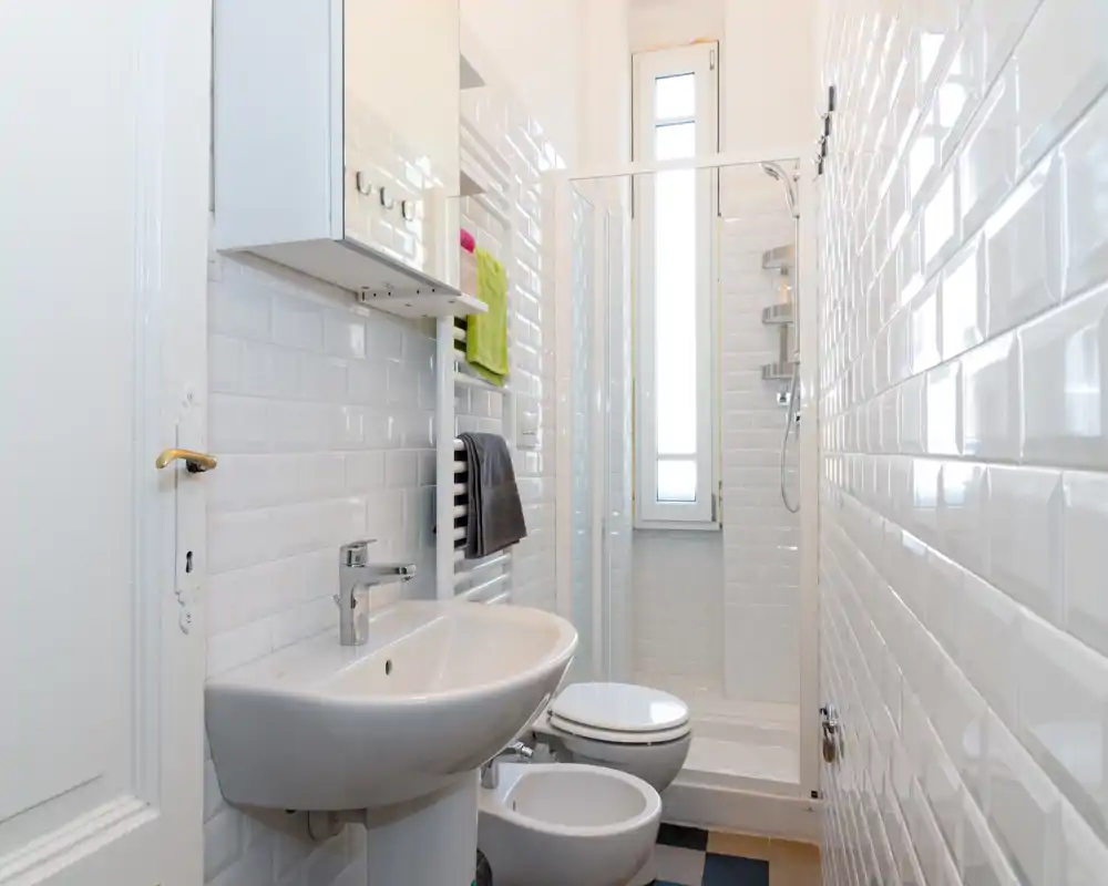 Baño pequeño y luminoso con azulejos blancos tipo metro, lavabo mural, bidé, inodoro y ducha con una ventana estrecha que aporta luz natural.