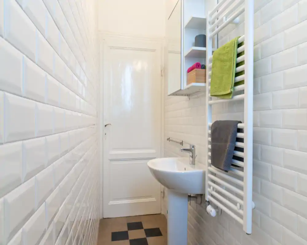 Pequeño baño luminoso con azulejos tipo metro blancos, lavabo de pedestal y toallero calefactado con toallas dobladas.