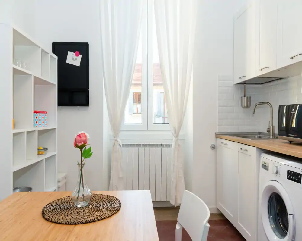 Cocina pequeña y luminosa con encimera de madera, lavadora integrada y una mesa pequeña con una flor que aporta calidez.