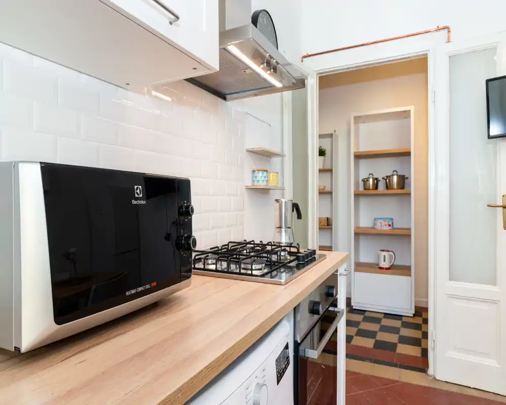 Cocina luminosa y compacta con cocina de gas, microondas sobre encimera de madera y vista a una despensa con estanterías — limpia, moderna y práctica para alquileres de corta estancia.